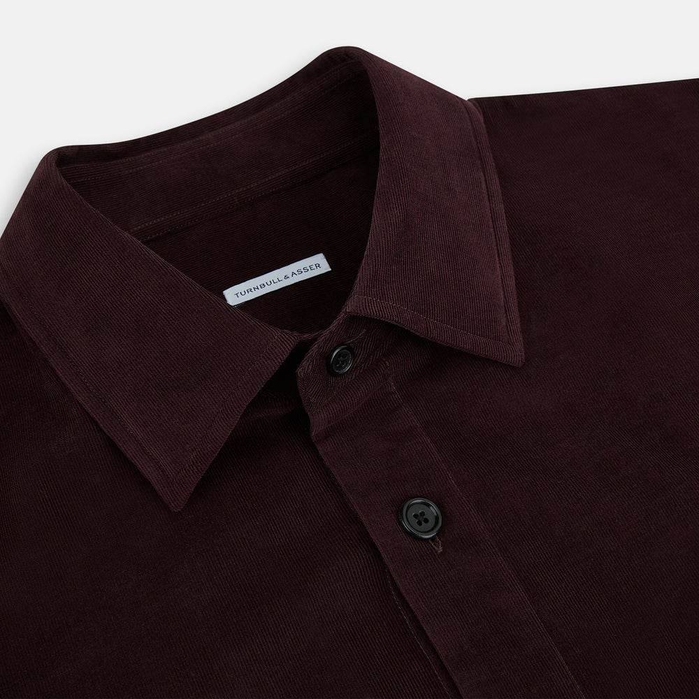 The Turnbull & Asser Burgundy Corduroy Hampstead Overshirt features long sleeves, a collar, a large left chest pocket, and two zippered side pockets, shown laid flat on a plain white background.