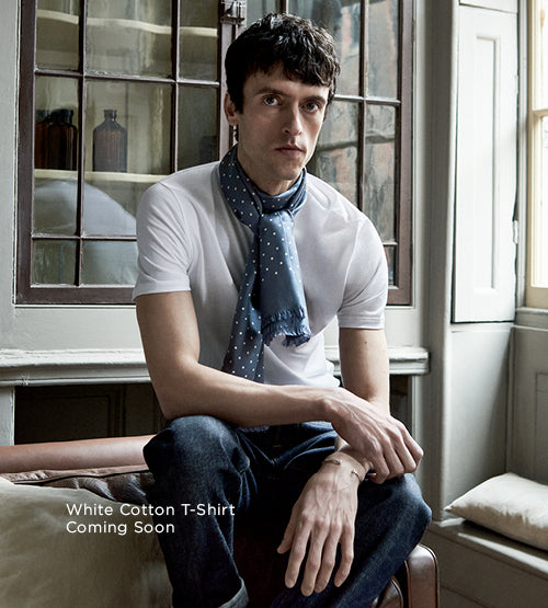 A man sits indoors wearing a white cotton T-shirt, blue jeans, and a blue polka dot scarf. Text on the image reads: White Cotton T-Shirt Coming Soon. A window and shelves are in the background.