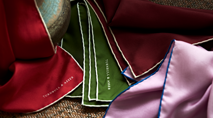 Colorful silk pocket squares—burgundy, green, and lavender—each trimmed in contrasting edges, are spread out on a woven surface. The green and burgundy squares are branded with TURNBULL & ASSER in white text.