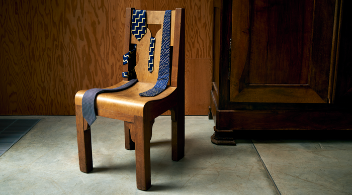 A wooden chair with a curved seat and backrest has several blue patterned neckties draped over it, set against a wooden wall and a dark wooden cabinet on a concrete floor.