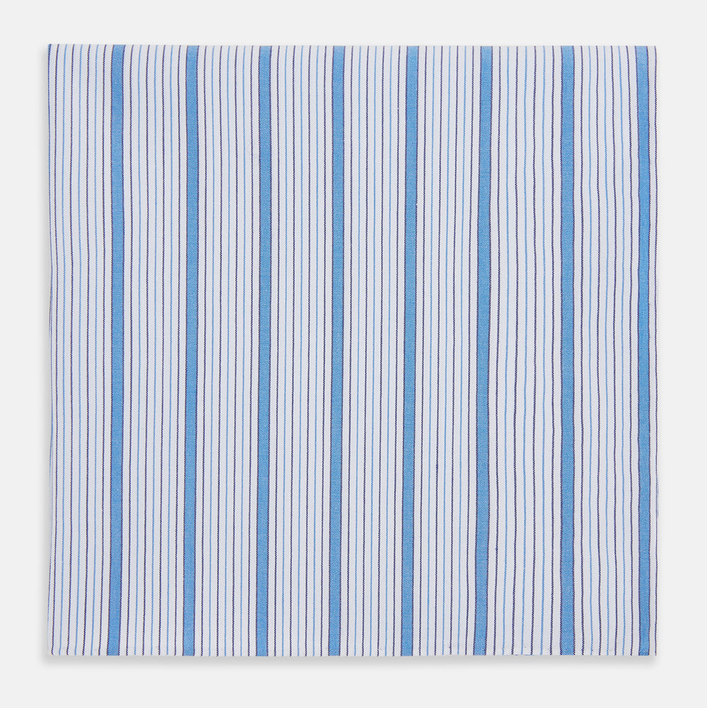 The Hand Rolled Blue Multi Track Stripe Handkerchief by Turnbull & Asser features a square white design with vertical light blue and navy stripes of varying widths—a timeless gentleman’s accessory.