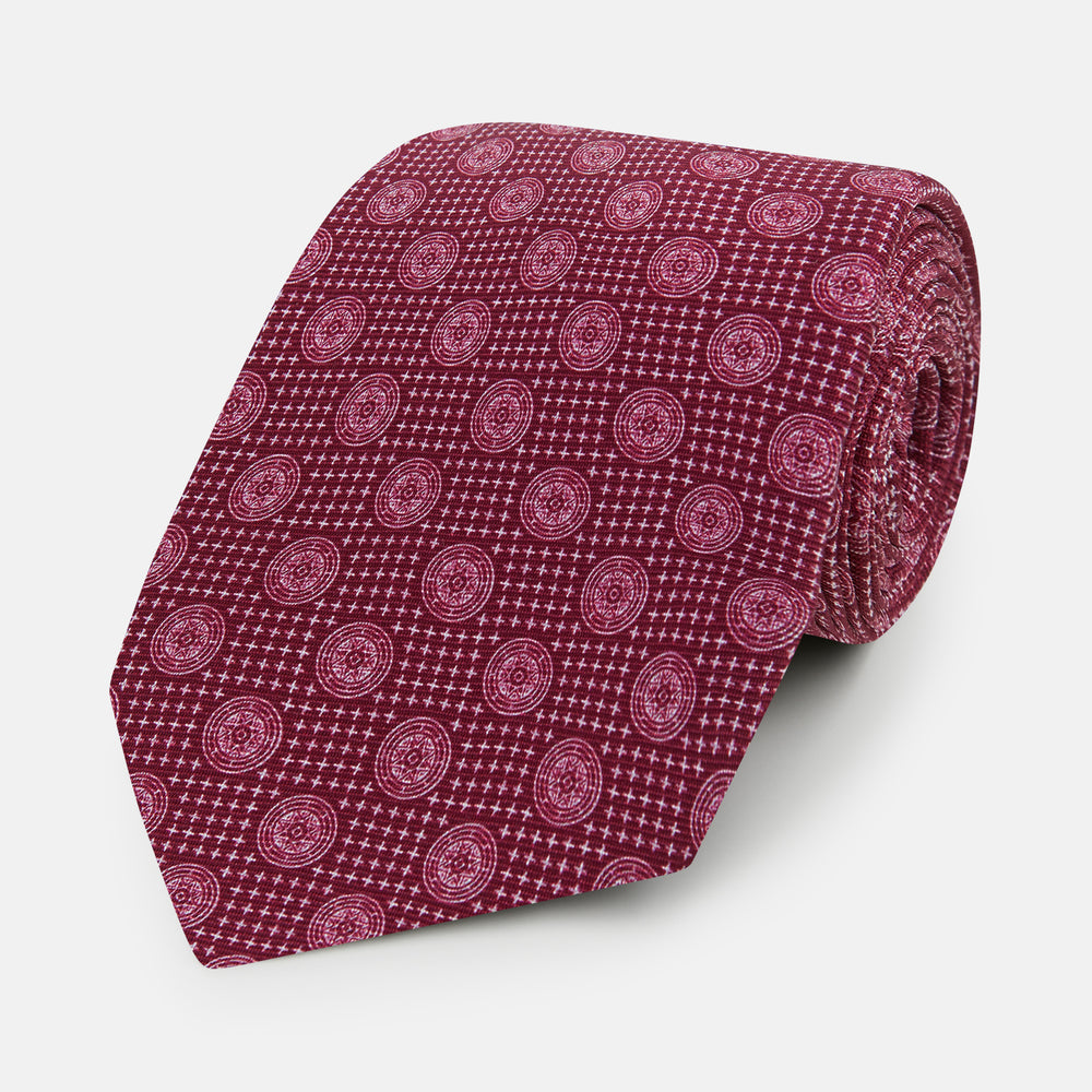 The Turnbull & Asser Burgundy Medallion Printed Silk Tie features small circular medallions and subtle light pink diagonal lines, crafted from fine English silk and displayed on a plain white background.