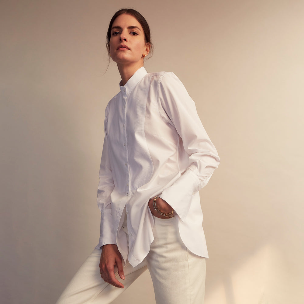 A person with curly hair wears the Turnbull and The Deck White Cotton Minimalist Shirt by Turnbull & Asser, paired with pants. Posed against a plain background, their minimalist silhouette is highlighted in this black and white image.