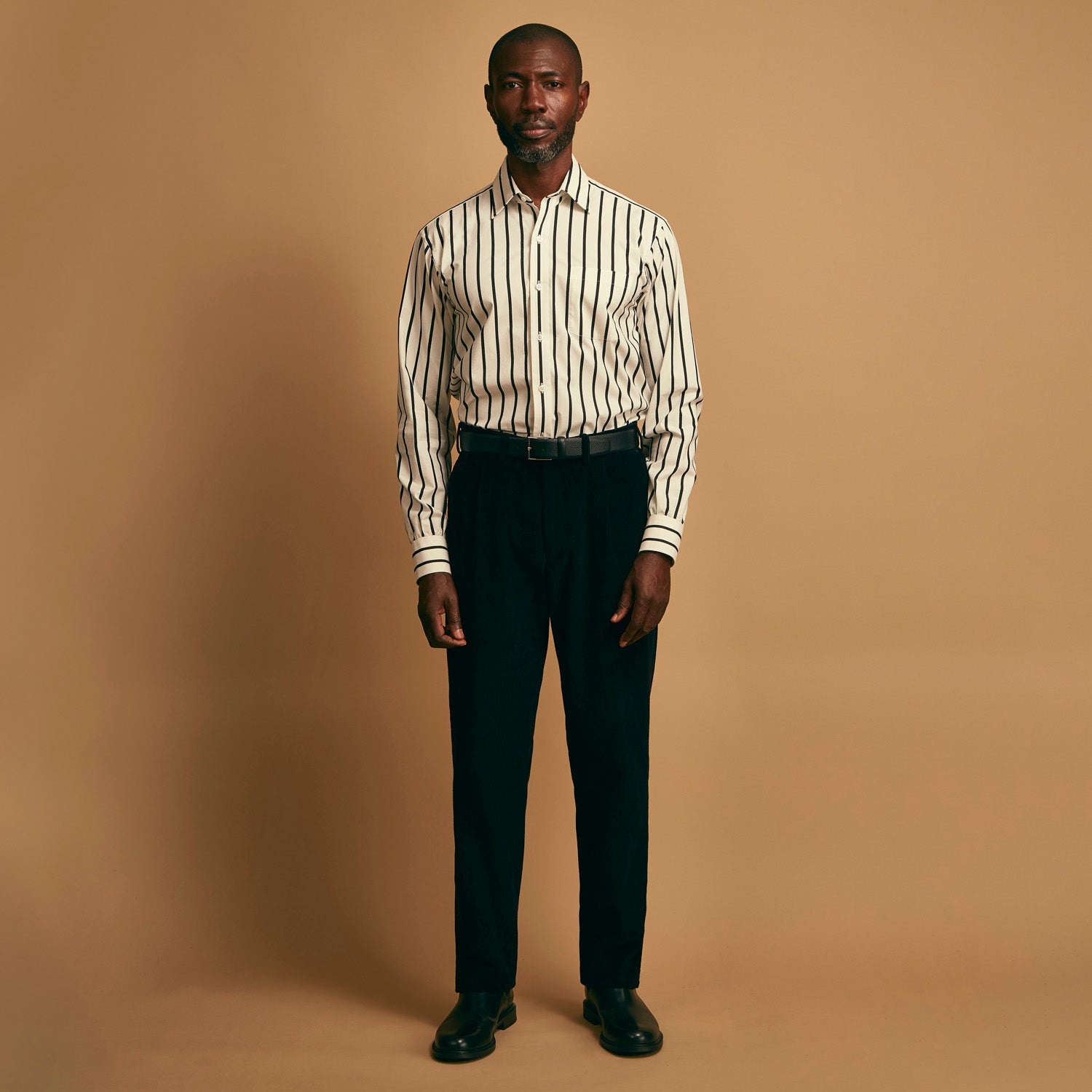 View 1 of product A man stands against a beige background, wearing a white striped shirt, Turnbull & Asser Black Cotton Cord Manson Trousers, and black shoes. He faces forward with a neutral expression.
