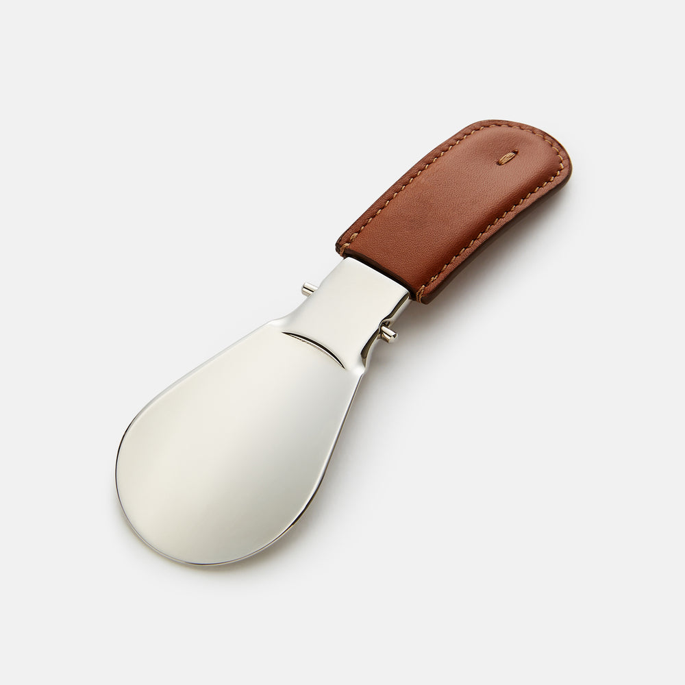 The Turnbull & Asser Camel Travel Shoe Horn features a wide, shiny silver blade and a brown Italian leather handle, displayed on a plain light background.