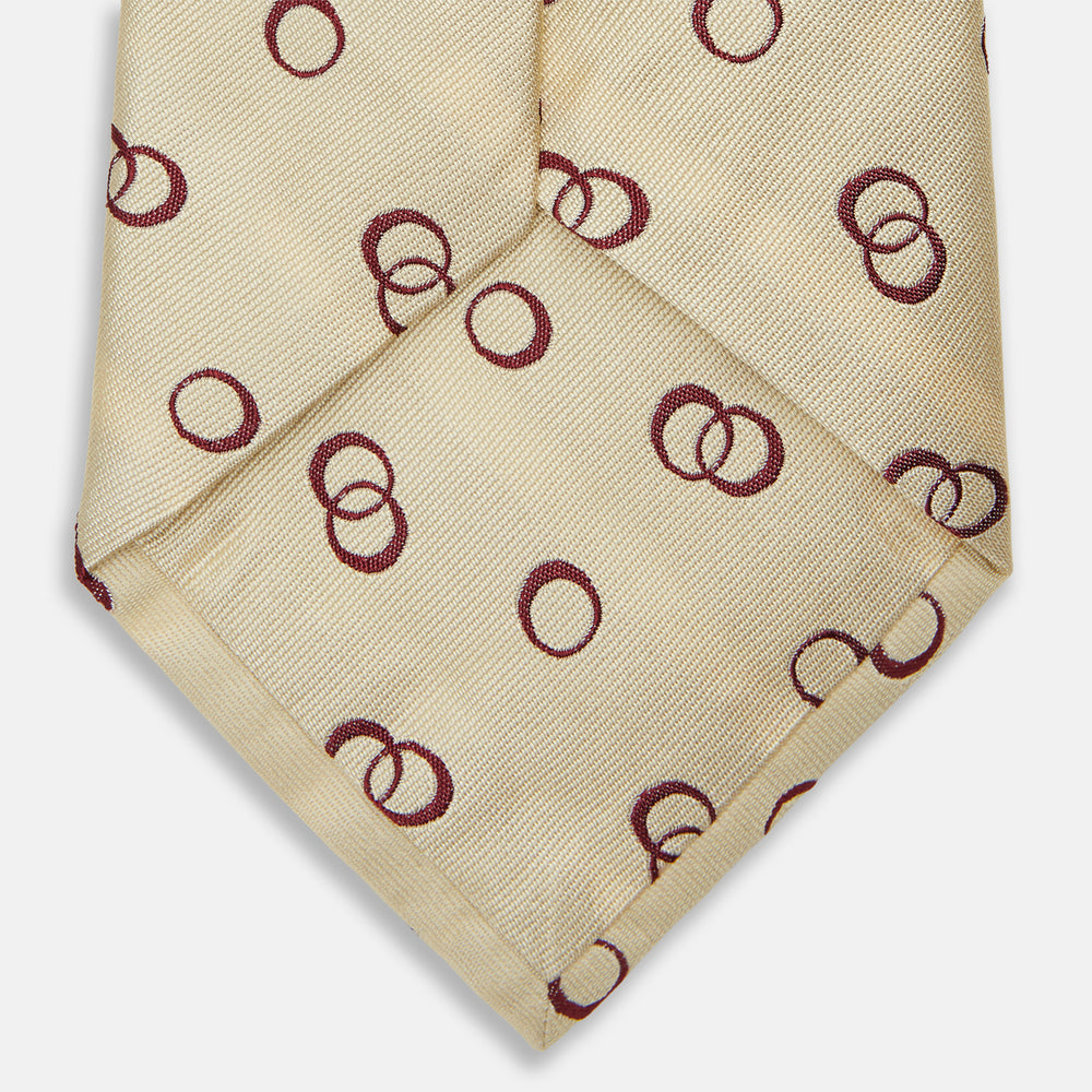 The Yellow Geometric Rings Silk Tie by Turnbull & Asser features a cream silk base with yellow geometric patterns and maroon rings, is hand-finished in England, and is shown rolled up on a white background.
