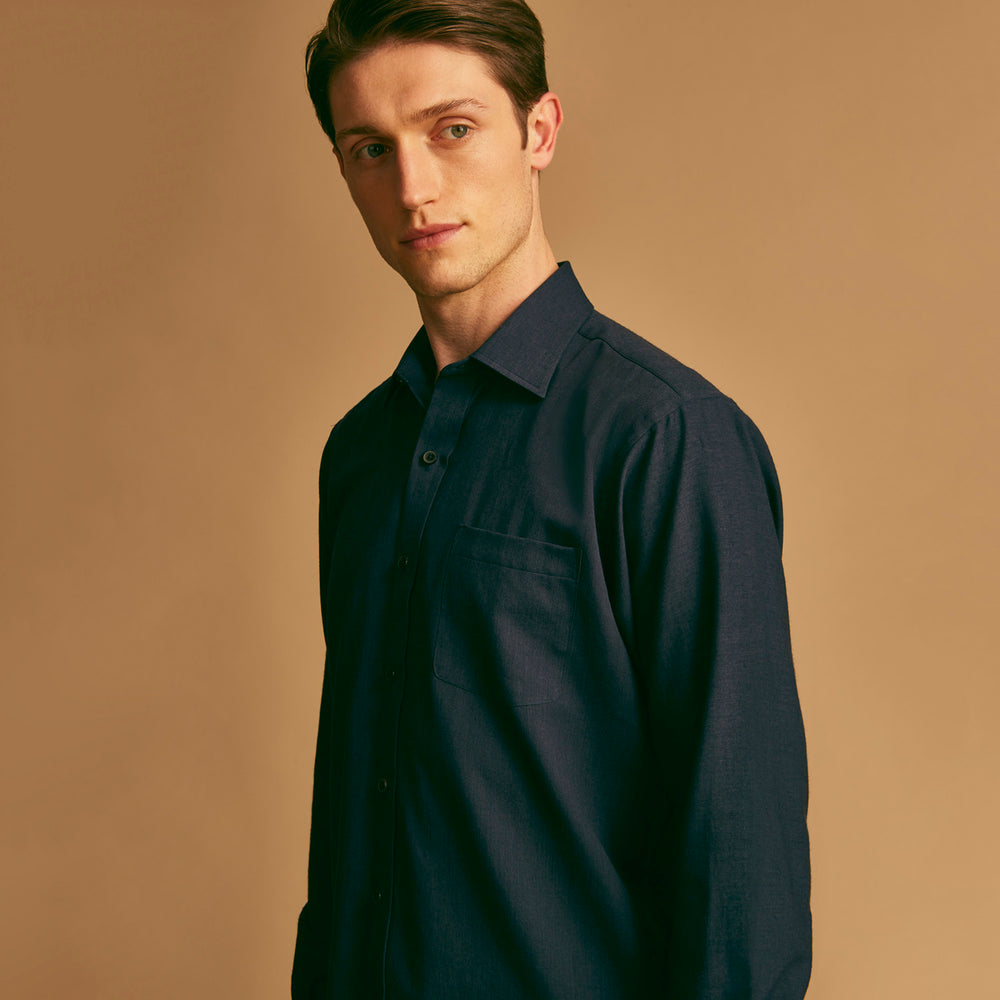A man stands against a beige background wearing the Turnbull & Asser Navy Weekend Fit Cotton & Wool Shirt with a Derby collar and 1 button cuffs, paired with brown pants and black shoes, looking to the side with a neutral expression.