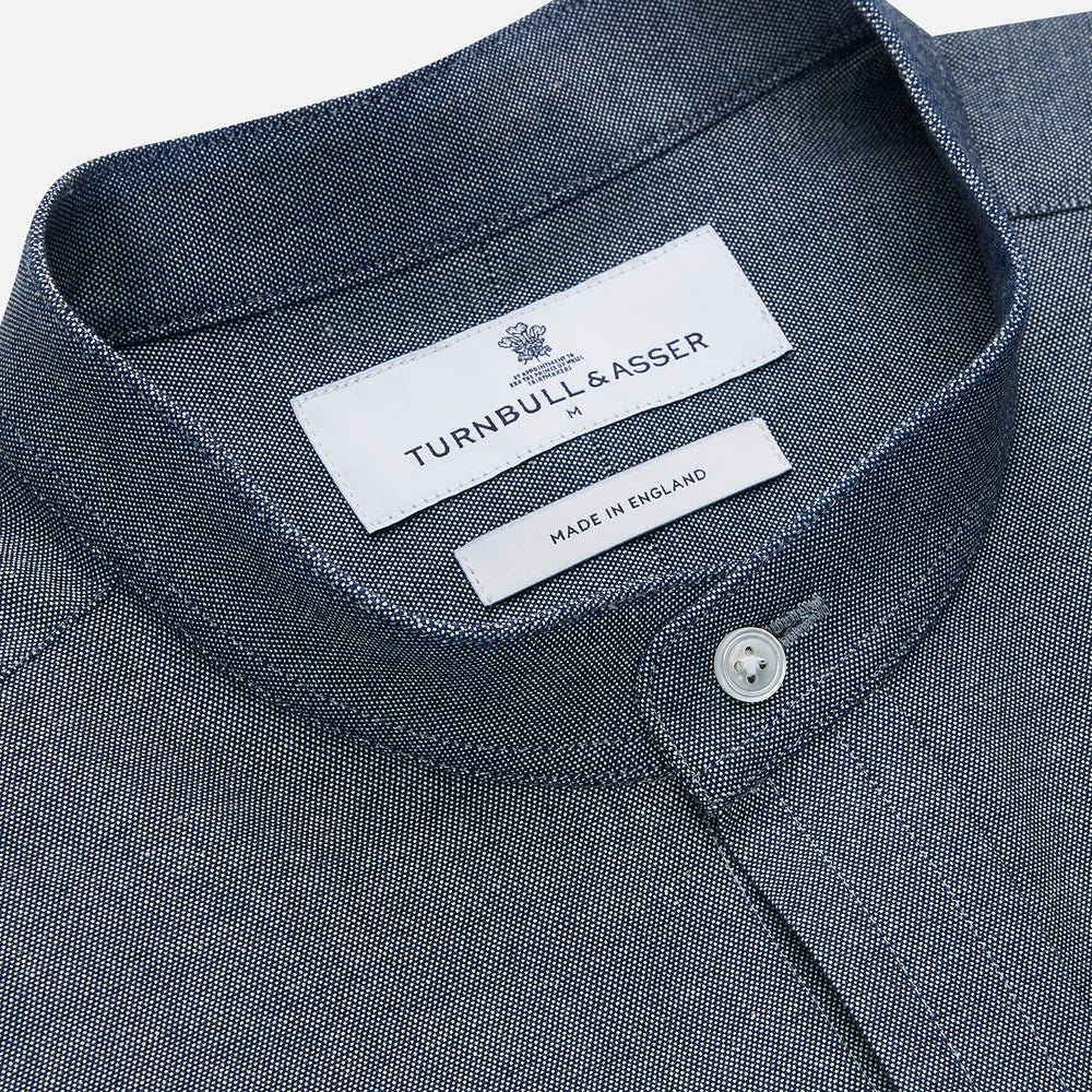 The Turnbull & Asser Plain Blue Oxford Cotton Weekend Fit Blake Shirt features long sleeves, a stand collar, and white buttons, displayed flat on a white background.
