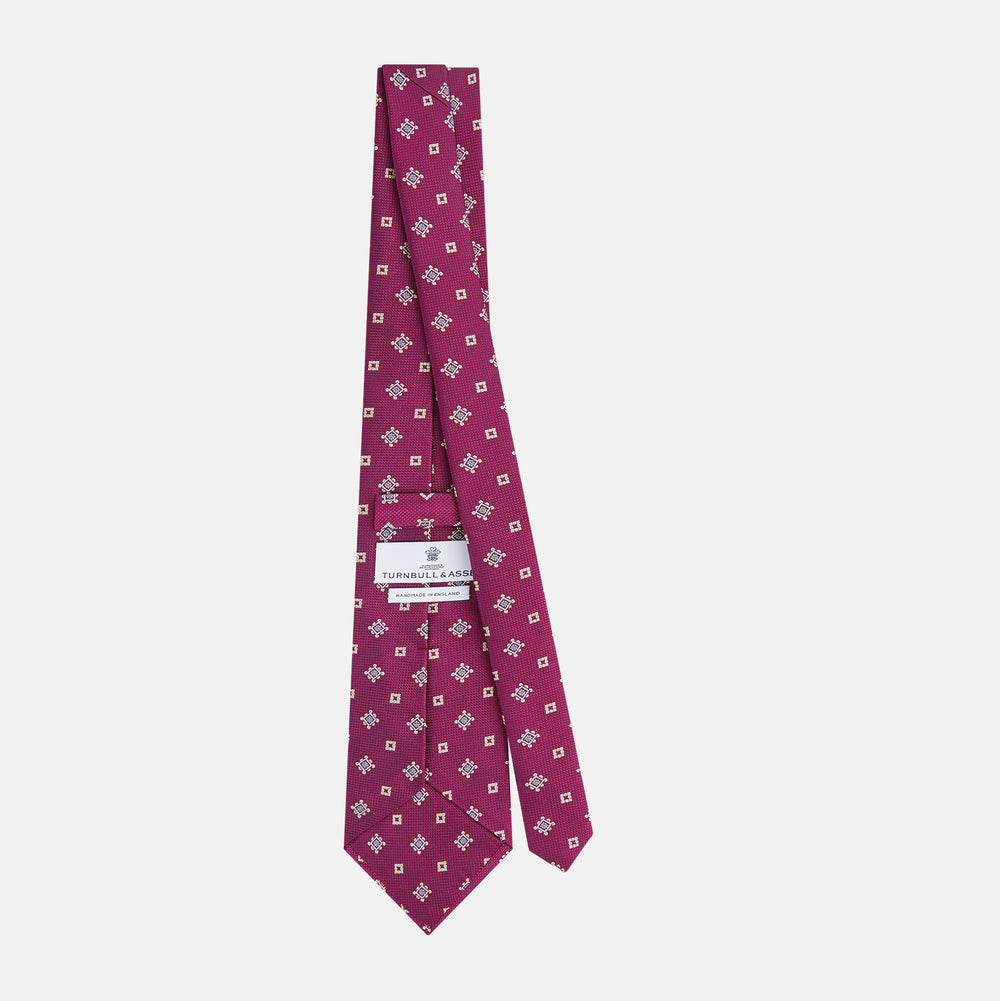 The Turnbull & Asser Purple Motif Silk Tie, rolled up and displayed on a light gray background, features small blue and white geometric designs—an elegant British touch for any formal event.