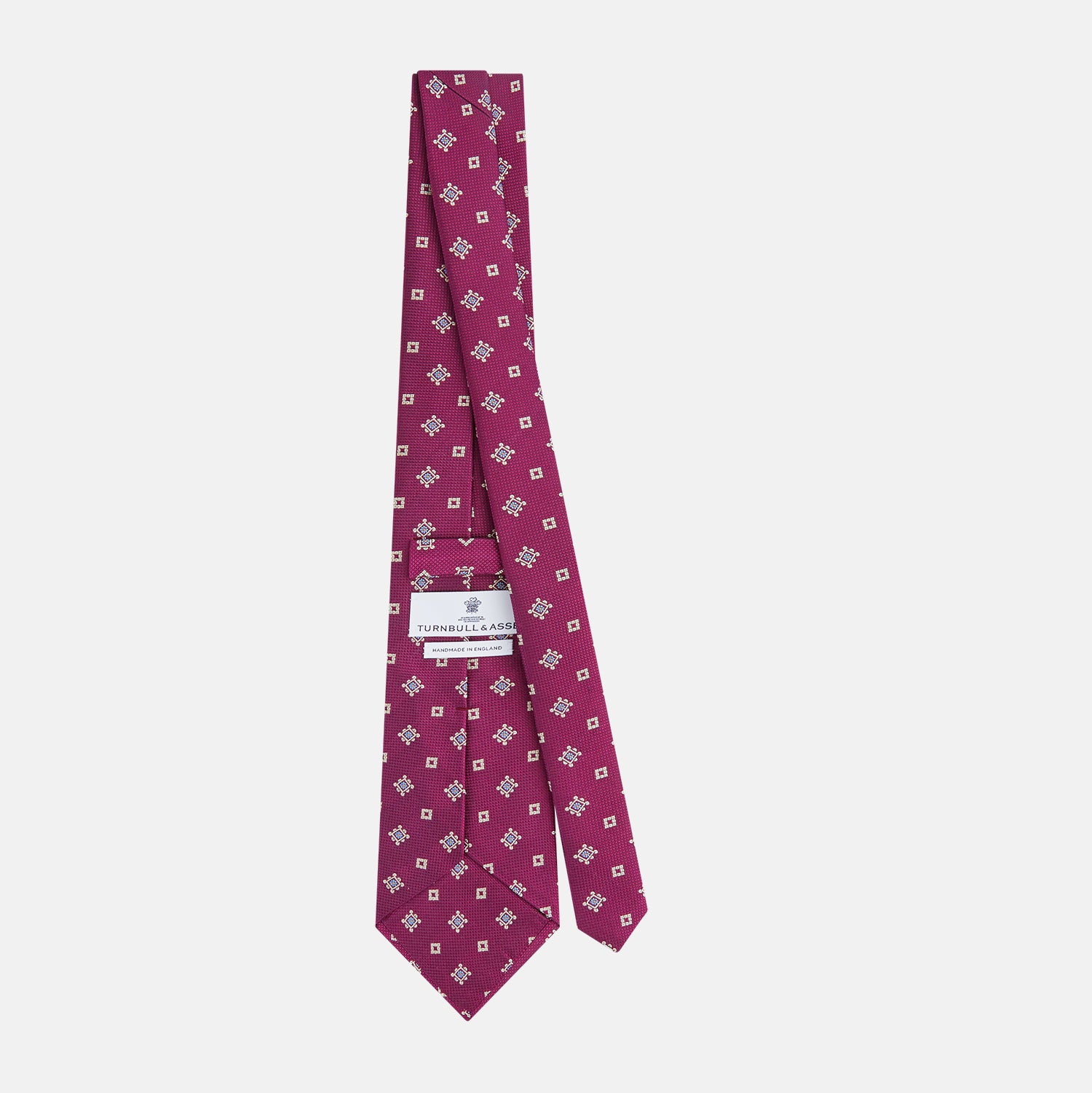 View 4 of product The Turnbull & Asser Purple Motif Silk Tie, rolled up and displayed on a light gray background, features small blue and white geometric designs—an elegant British touch for any formal event.