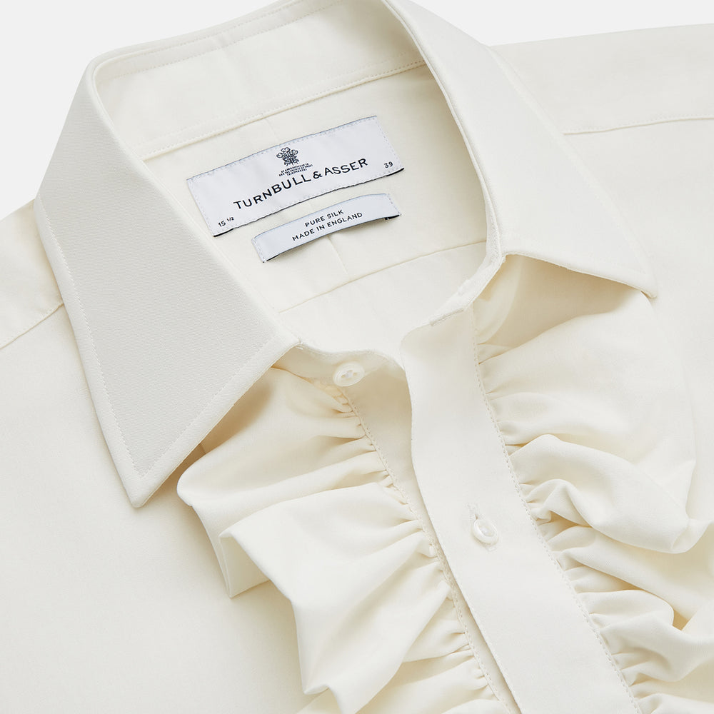A man in a Cream Silk Regular Fit Ruffle-Front Wilfred Shirt by Turnbull & Asser stands against a dark background, one hand on a chair, gazing thoughtfully to the side.