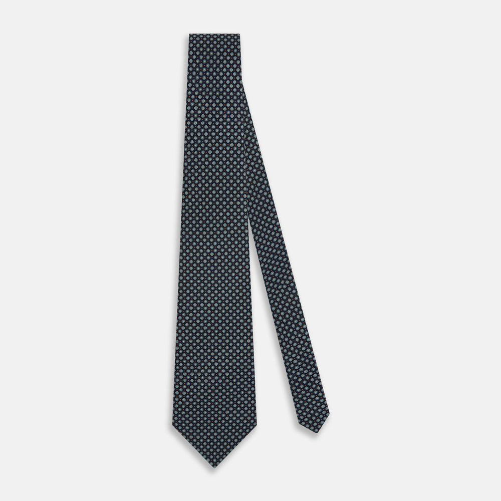 The Turnbull & Asser Navy Geometric Floral Silk Tie features a dark blue background with a repeating small geometric pattern in light blue, yellow, and red, shown rolled up on a plain white surface.