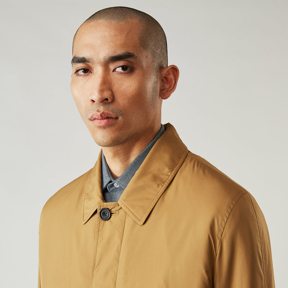 A person wears the Turnbull & Asser Camel Quintern Sealup Raincoat with Detachable Liner, paired with dark pants, standing against a plain light gray background, showing the sleek technical rainwear style with one hand in a pocket.