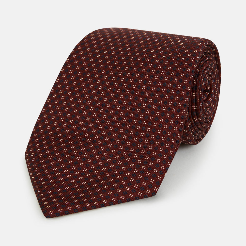 The Burgundy Geometric Motif Silk Tie by Turnbull & Asser features blue and white dots and is displayed neatly rolled on a plain white background.