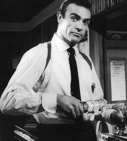 A man in a white dress shirt, tie, and suspenders stands indoors, holding a cocktail shaker and looking toward the camera with a slight smile in a black-and-white photo.
