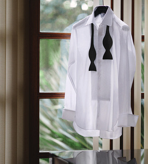 A white dress shirt with a black bow tie draped around the collar hangs on a hanger in front of a sunlit window with green foliage visible outside.