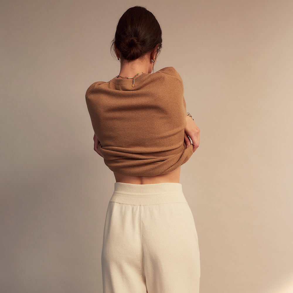 A woman stands barefoot indoors against a neutral wall, wearing Turnbull & Asser Women's Cream Cashmere Knitted Tabitha Bottoms and a light brown sweater. Soft sunlight casts a gentle shadow on the wall beside her.