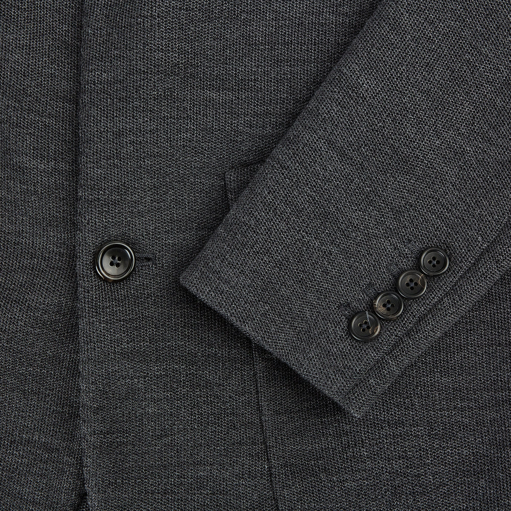 The Turnbull & Asser Graphite Knitted Oxford Blazer for men features a dark gray hue, two buttons, notched lapels, and three front pockets—a modern British-inspired piece styled elegantly against a light background.