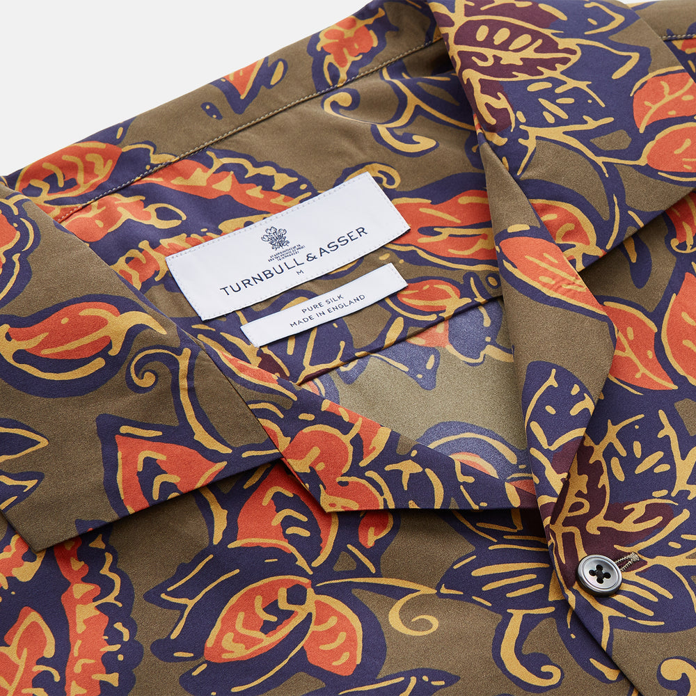 The Turnbull & Asser Khaki Green Floral Silk Holiday Fit Winnington Shirt features a bold floral print with orange, yellow, and purple on an olive background, plus a spread collar and chest pocket for versatile style.