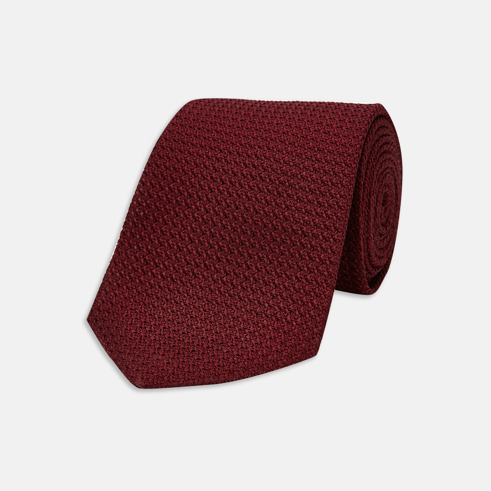 The Turnbull & Asser 7 fold Grenadine Silk Plain Tie is shown rolled on a white background, highlighting its textured maroon design as a versatile addition to any wardrobe.