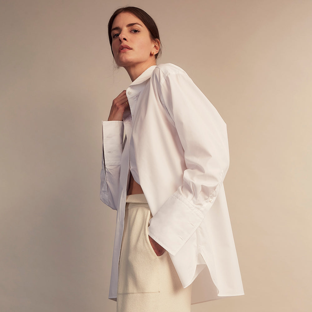 A woman with straight brown hair pulled back wears the Turnbull & Asser White Cotton Marlene Shirt, featuring classic menswear details, paired with high-waisted cream trousers against a neutral background.
