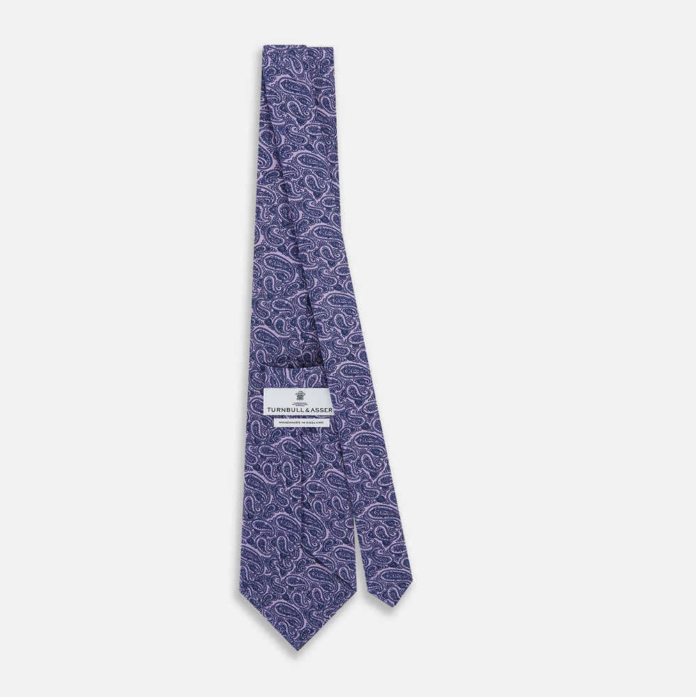 The Turnbull & Asser Lilac Paisley Silk Tie is shown rolled up, featuring a blue paisley pattern on a light purple background and displayed on a white surface.