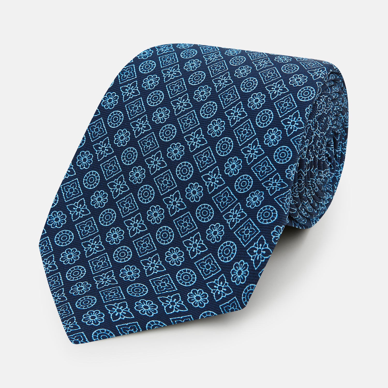 View 1 of product The Turnbull & Asser Dark Navy Floral Tile Pattern Printed Silk Tie, rolled up and displayed on a white background, features a blue geometric and floral pattern.