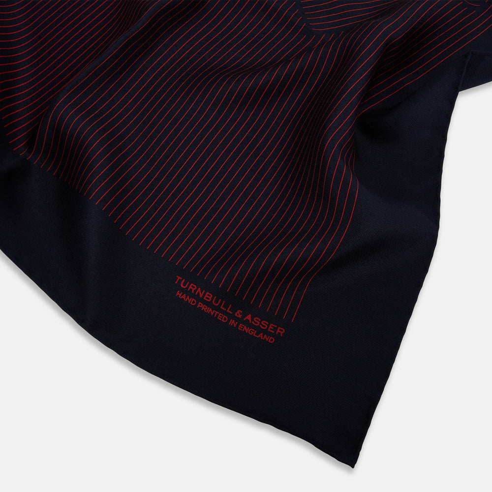 The Turnbull & Asser Navy and Red Stripe Silk Pocket Square features thin red stripes on navy silk, with "HAND PRINTED IN ENGLAND" in red at the corner. A folded corner shows its hand-rolled edges and refined design.