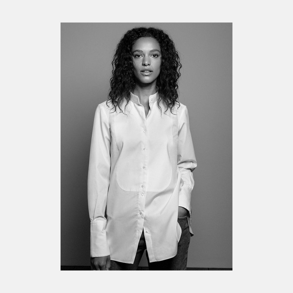 A person with curly hair wears the Turnbull and The Deck White Cotton Minimalist Shirt by Turnbull & Asser, paired with pants. Posed against a plain background, their minimalist silhouette is highlighted in this black and white image.