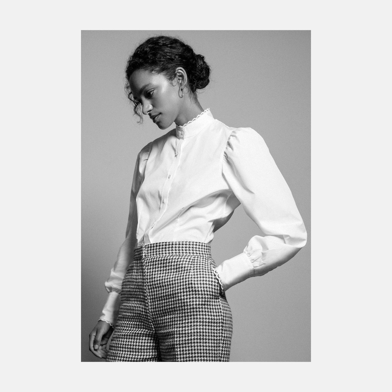 View 1 of product A woman stands in profile, looking down with hands in her pockets, wearing the Turnbull & Asser Turnbull and The Deck White Cotton Expressionist Shirt, paired with high-waisted black-and-white houndstooth pants.