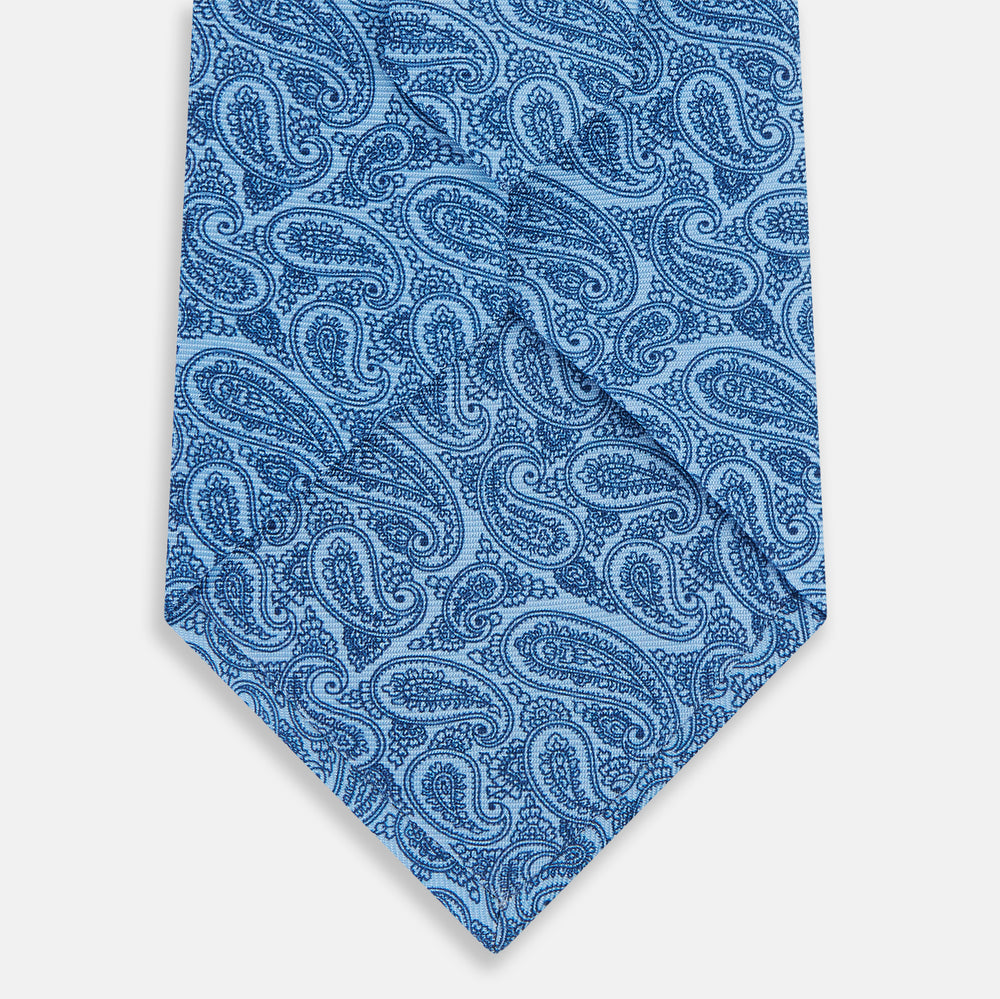 The Turnbull & Asser Blue Paisley Printed Silk Tie features a blue paisley pattern on a light blue background and is shown rolled up on a plain white surface.