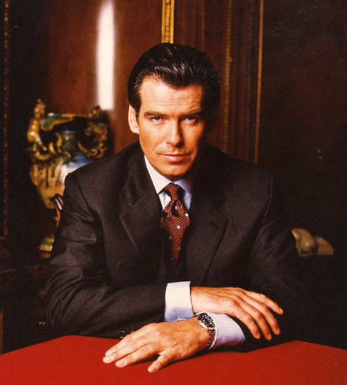 A man in a dark suit and patterned tie sits at a table with his hands folded, looking directly at the camera, against an elegant, wood-paneled background with ornate decor.