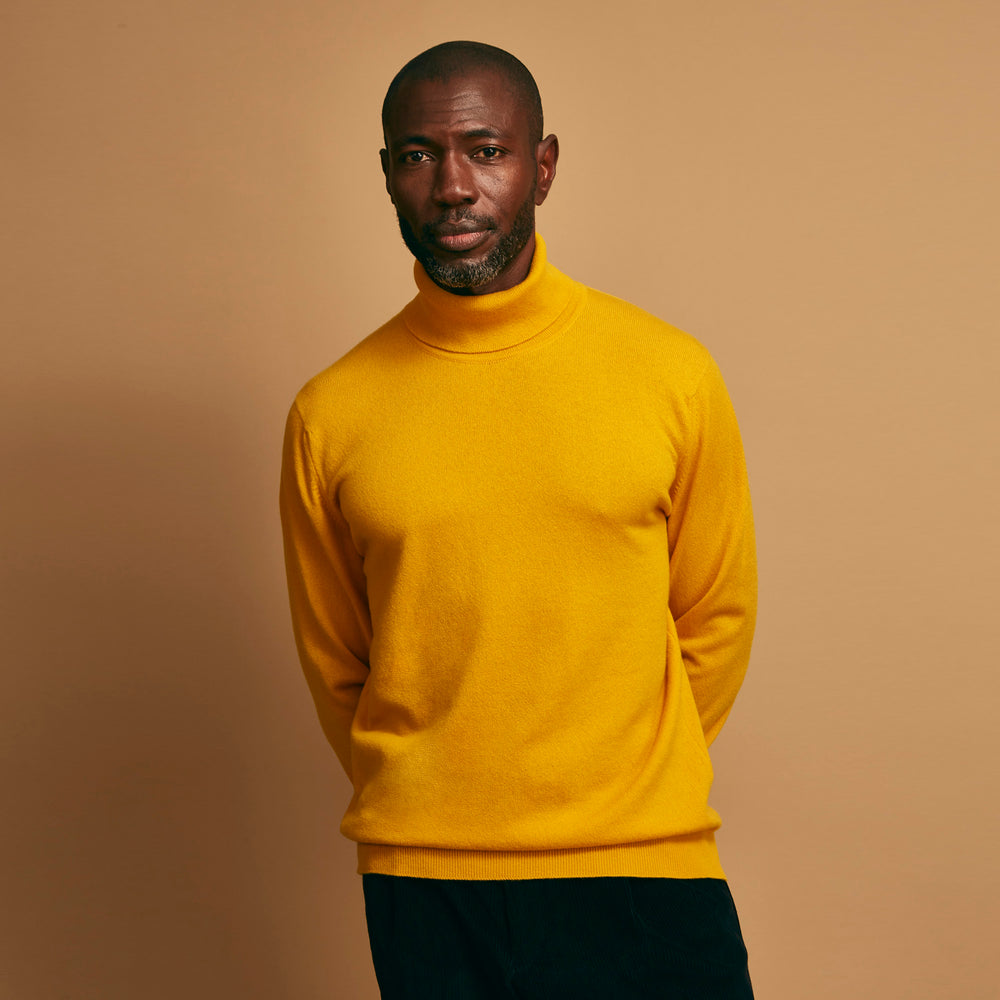 A man with short hair and a beard wears the Gold Aubrey Cashmere Roll Neck Jumper by Turnbull & Asser with dark pants, standing against a plain beige background and calmly looking at the camera, highlighting this stylish autumn sweater look.