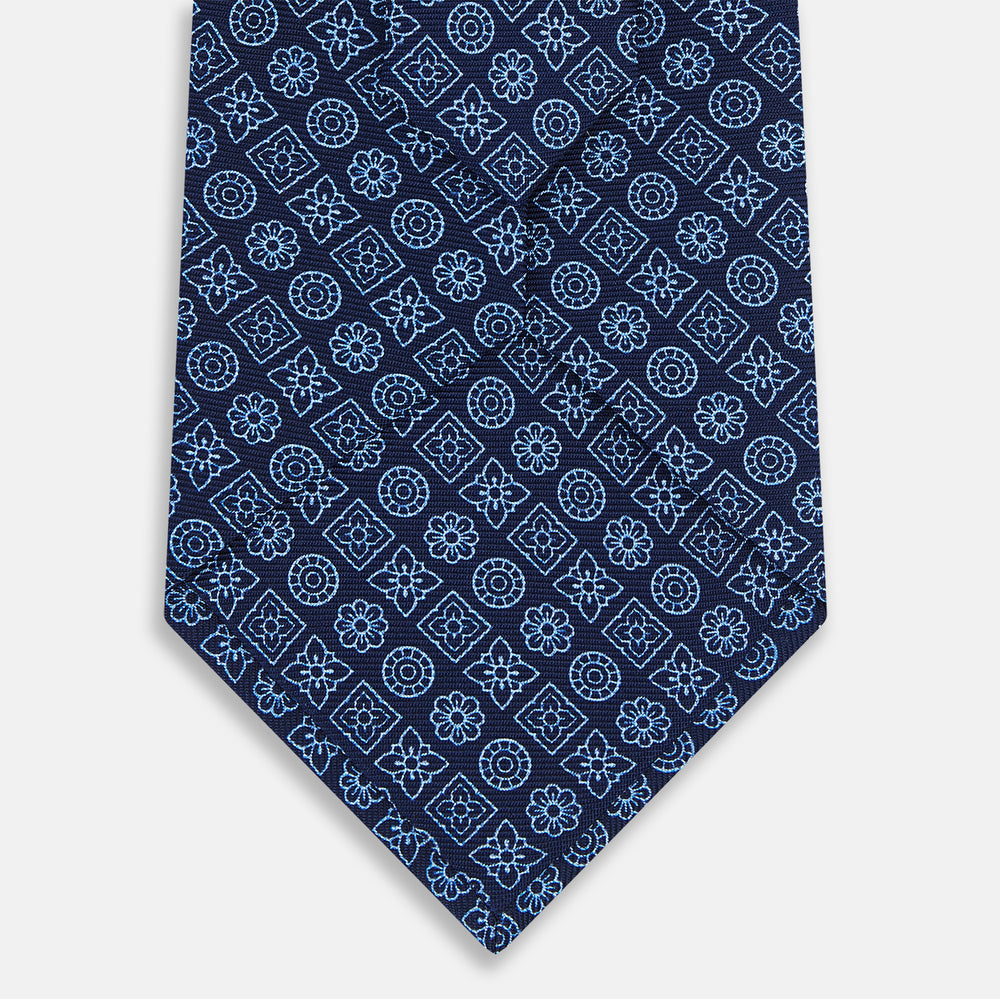 The Turnbull & Asser Dark Navy Floral Tile Pattern Printed Silk Tie, rolled up and displayed on a white background, features a blue geometric and floral pattern.