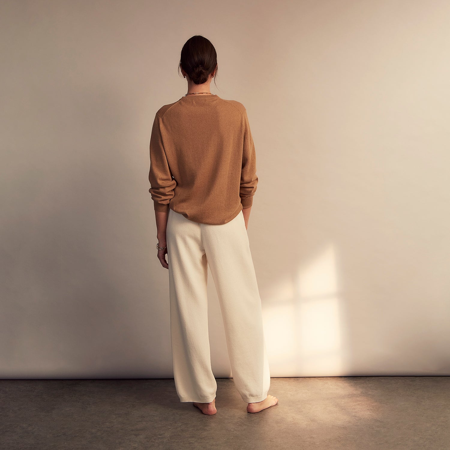 View 3 of product A woman stands barefoot indoors against a neutral wall, wearing Turnbull & Asser Women's Cream Cashmere Knitted Tabitha Bottoms and a light brown sweater. Soft sunlight casts a gentle shadow on the wall beside her.