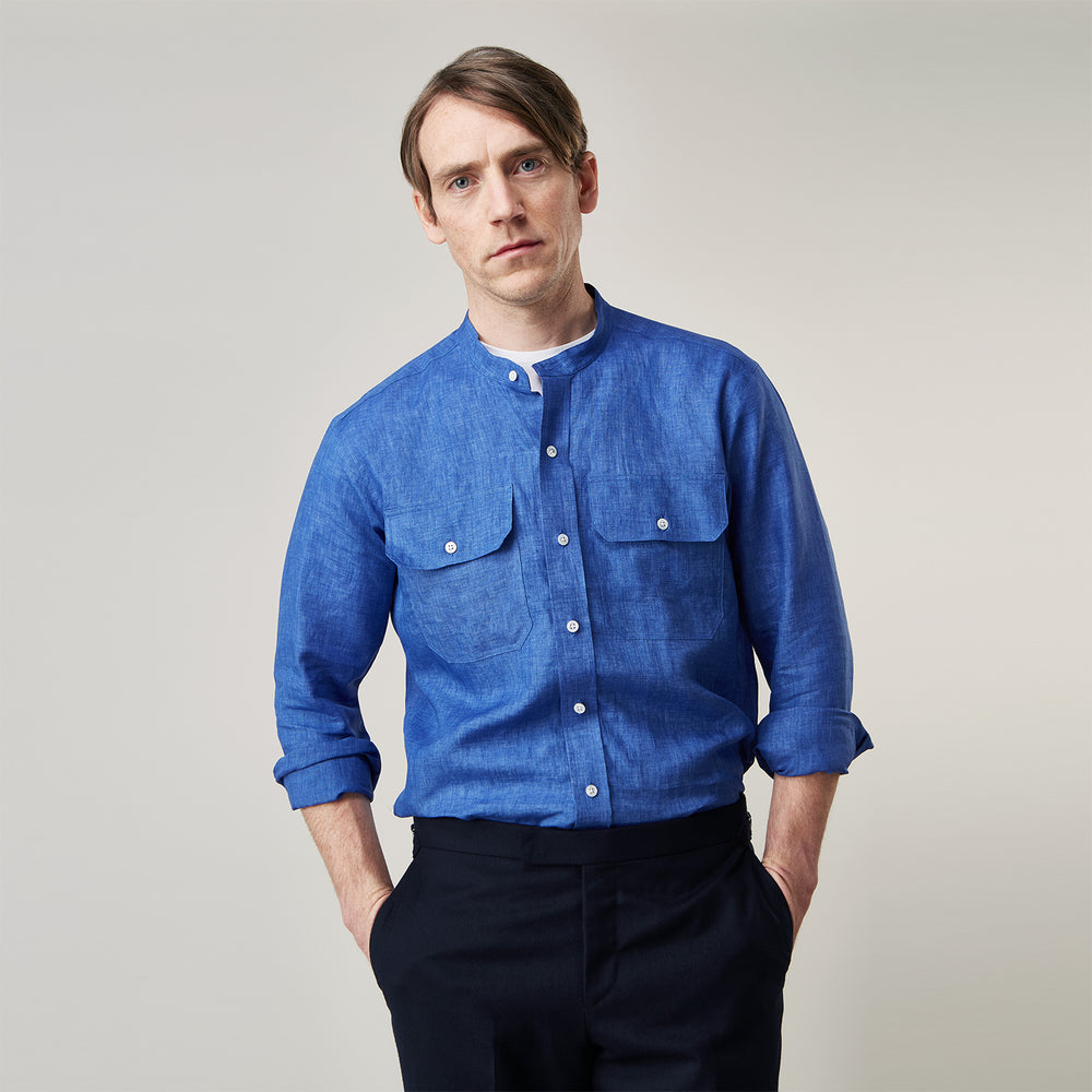 A man faces forward with a neutral expression, hands in his pockets, wearing the Turnbull & Asser Plain Indigo Linen Weekend Fit Shirt with a stand collar, styled with dark pants and black shoes against a plain light background.