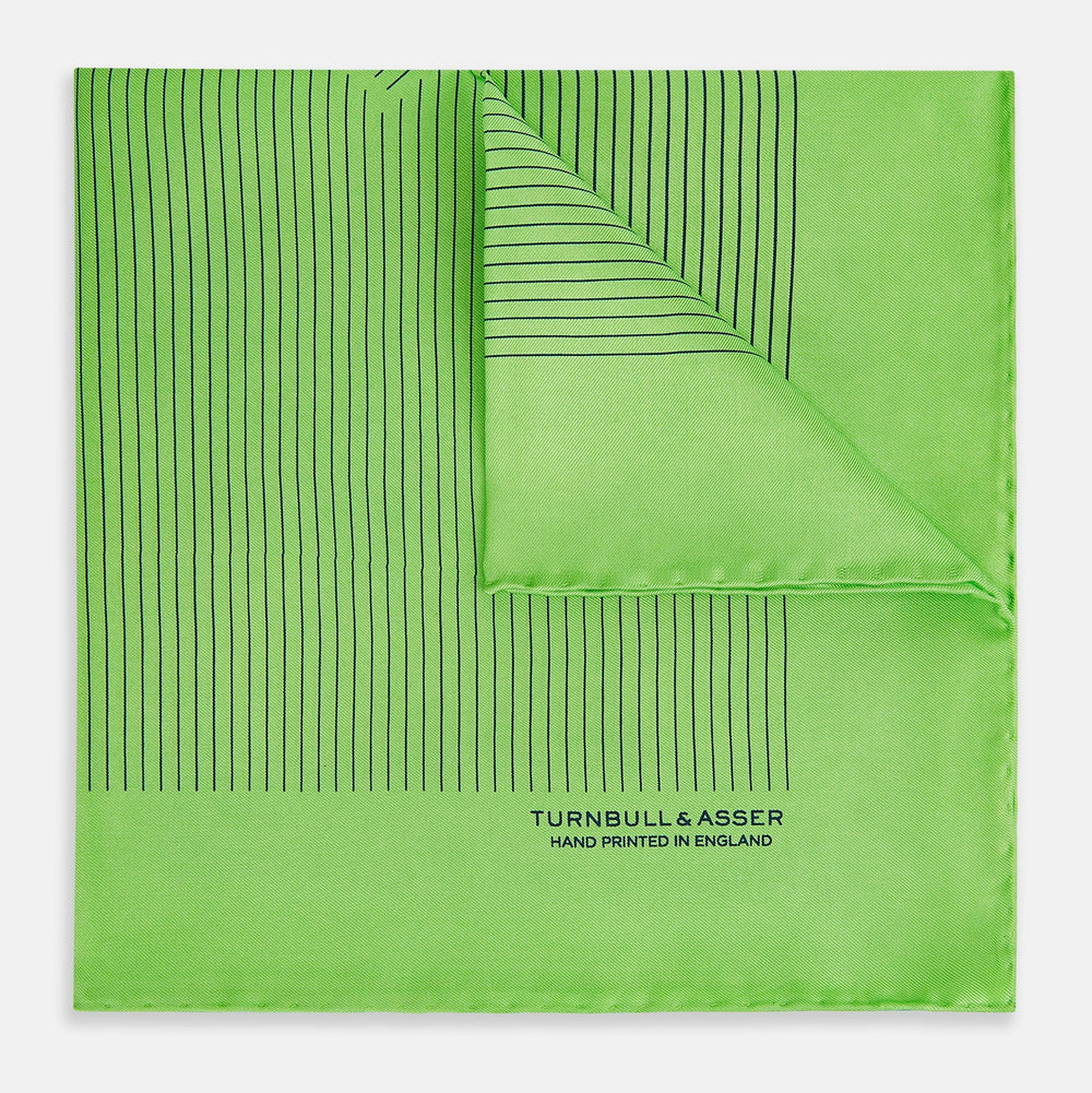 The Fern Green Stripe Silk Pocket Square by Turnbull & Asser features thin black vertical stripes, celebrates British heritage, and is inscribed “TURNBULL & ASSER HAND PRINTED IN ENGLAND” in one corner; one corner is folded inward.