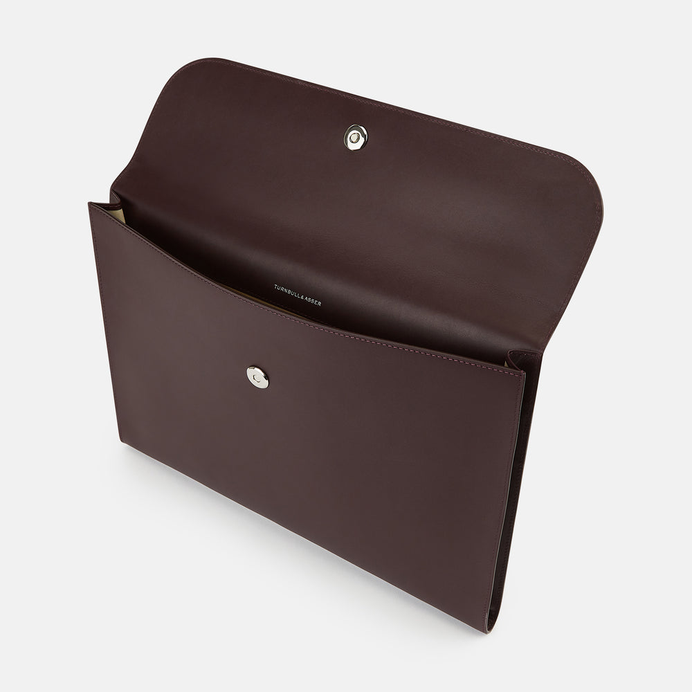 The Turnbull & Asser Burgundy Leather Snap-Fasten Pouch is a sleek, envelope-style laptop accessory with a smooth finish, curved flap, and elegant snap closure, ideal for business travel against a white background.