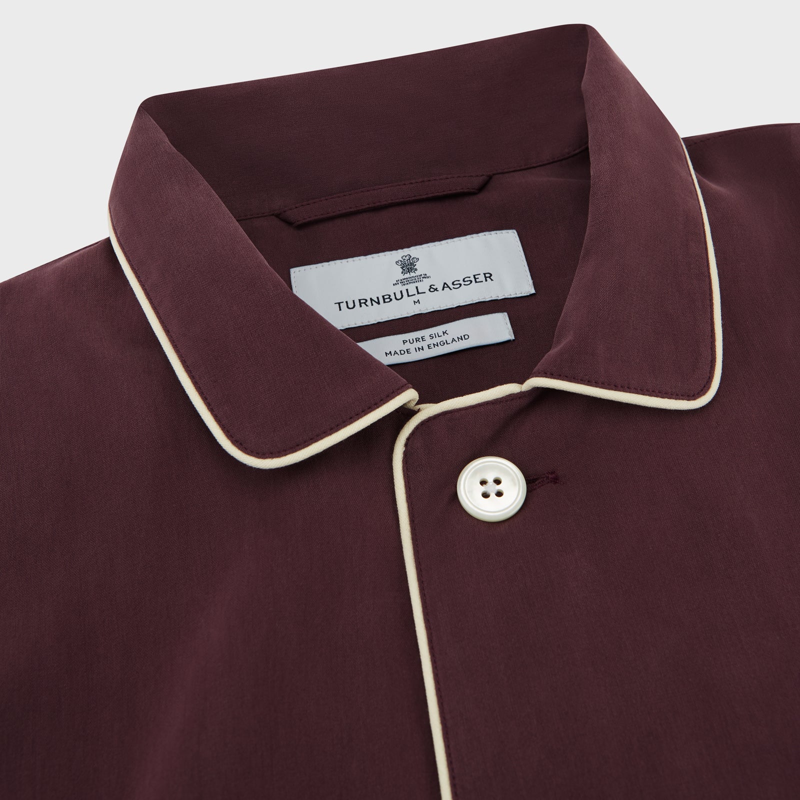 View 2 of product The Turnbull & Asser Plain Plum Silk Modern Pyjama Set, made in England, features white piping, a button-up long-sleeve top with three pockets, and matching pants. The pyjamas are shown against a white background.