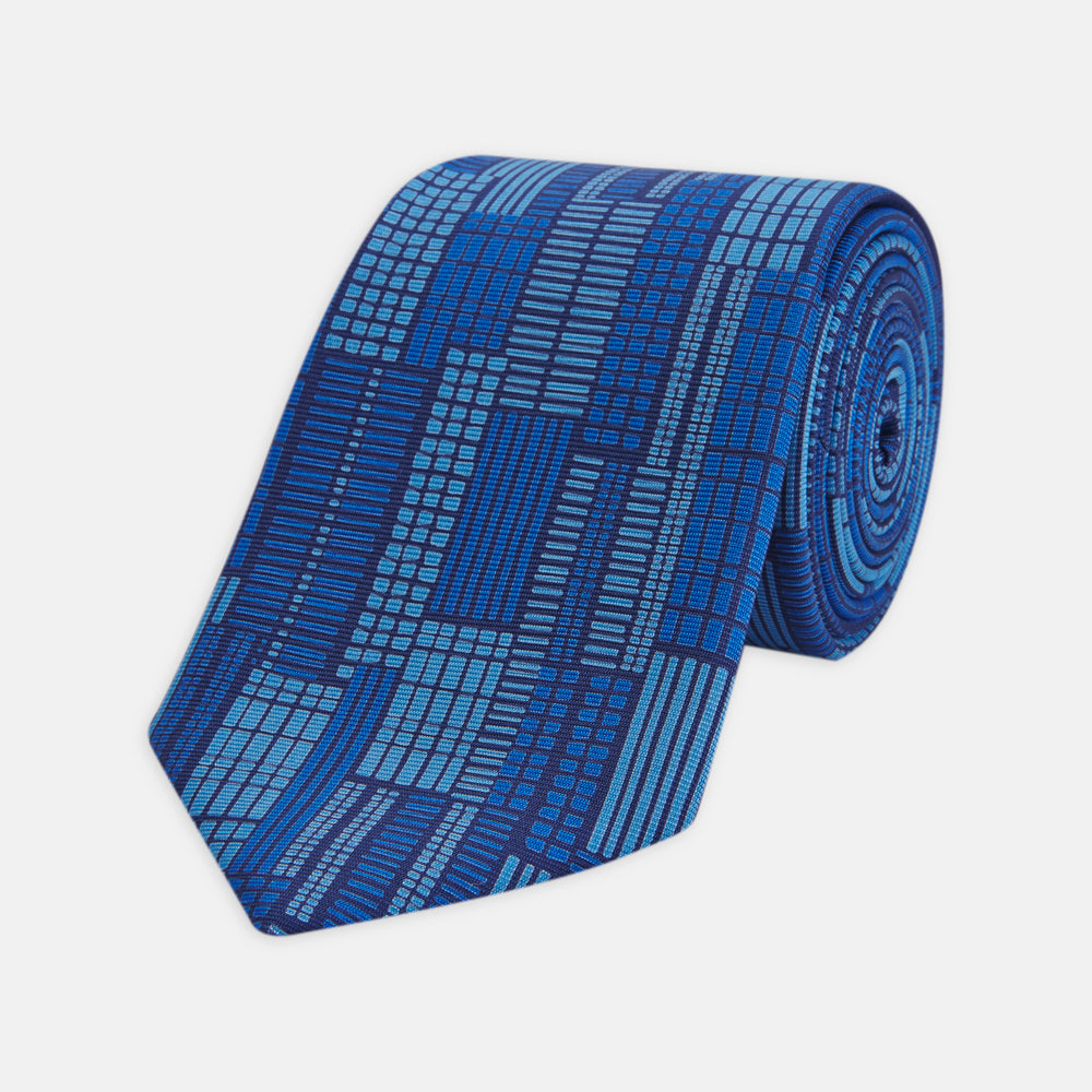 The Turnbull & Asser Blue Motif Printed Foulard Silk Tie showcases a geometric pattern in varying blue shades, making it a striking and essential accessory for any wardrobe.