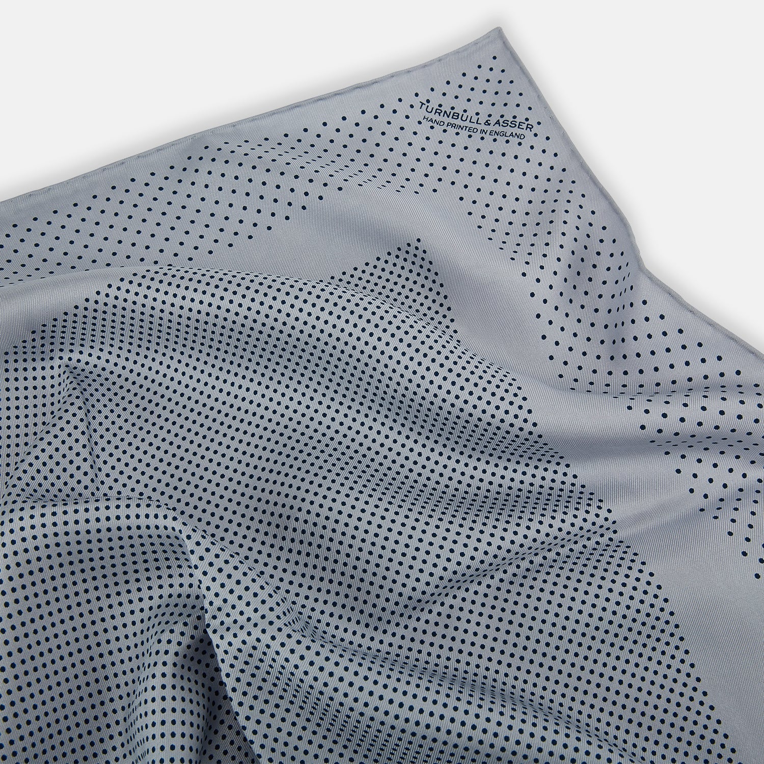 View 7 of product The Turnbull & Asser Grey Dot Silk Pocket Square features a light gray silk design with navy polka dots and a solid border. Elegantly handmade, it is shown with one corner folded to display both sides.