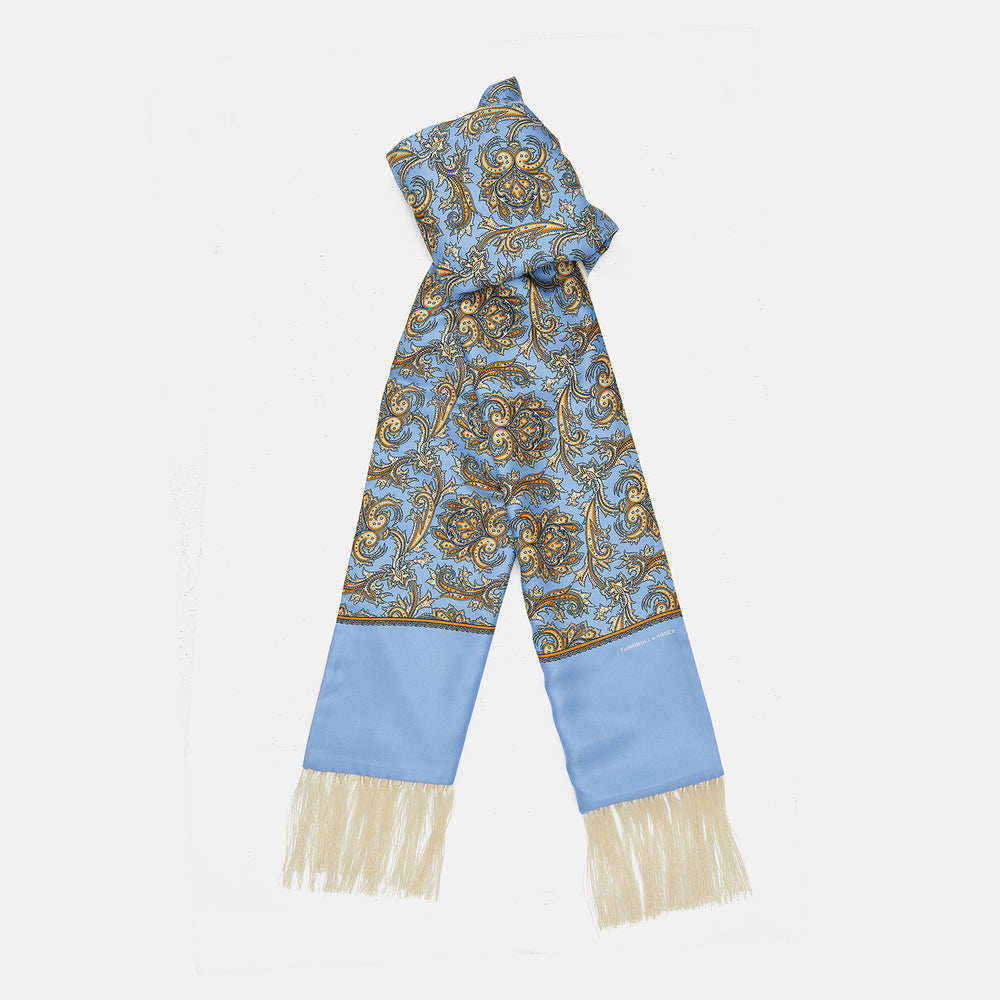 The Turnbull & Asser GGSC3128 5665 06 OS is a blue paisley silk scarf with ornate gold designs, solid light blue ends, and cream fringes. Shown laid flat with one end folded, it highlights its luxurious style.