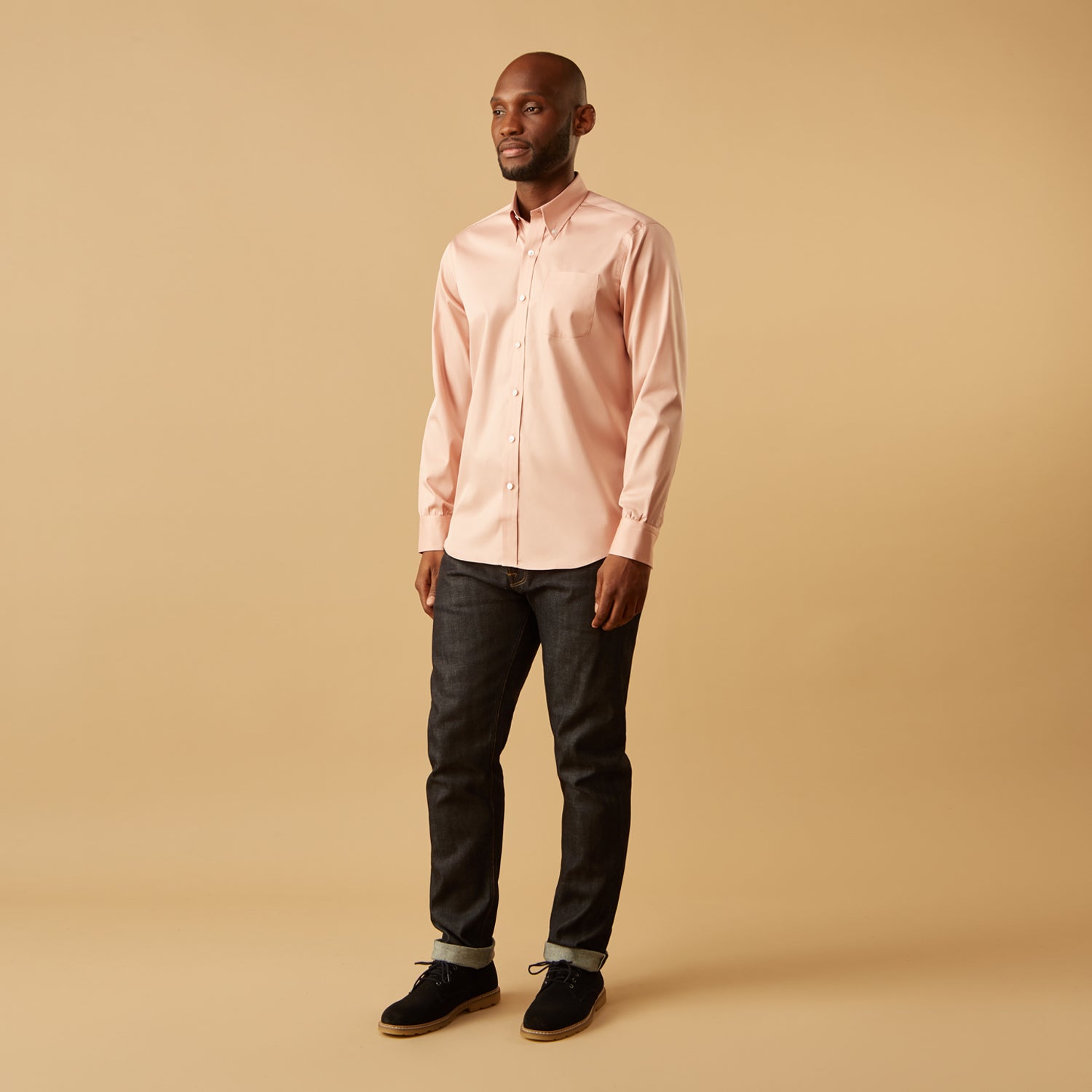 View 1 of product A man faces forward in a relaxed pose against a beige background, wearing the Turnbull & Asser Pink Piece Dyed Weekend Fit Suffolk Shirt with a button-down collar, dark cuffed jeans, and black shoes.