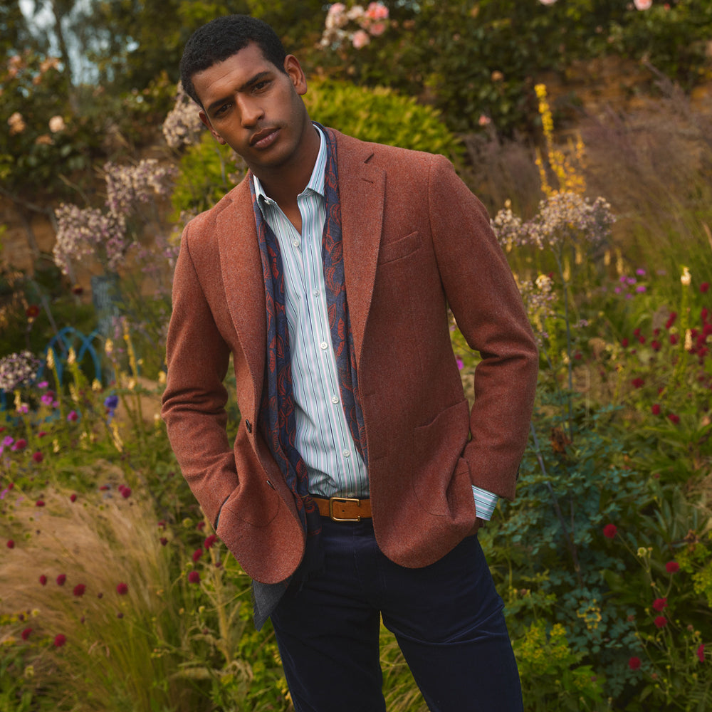 A man stands in a lush garden, hands in pockets and relaxed, wearing the Turnbull & Asser Red Lambswool Herringbone Bern Blazer, a striped dress shirt, patterned scarf, and dark trousers.