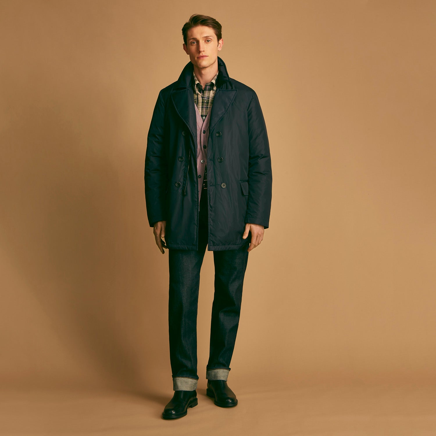 View 1 of product A man stands against a beige background, wearing the Turnbull & Asser Navy Burrows Double Breasted Coat over a plaid shirt and mauve cardigan, paired with dark rolled-up jeans and black shoes, looking directly at the camera with a neutral expression.