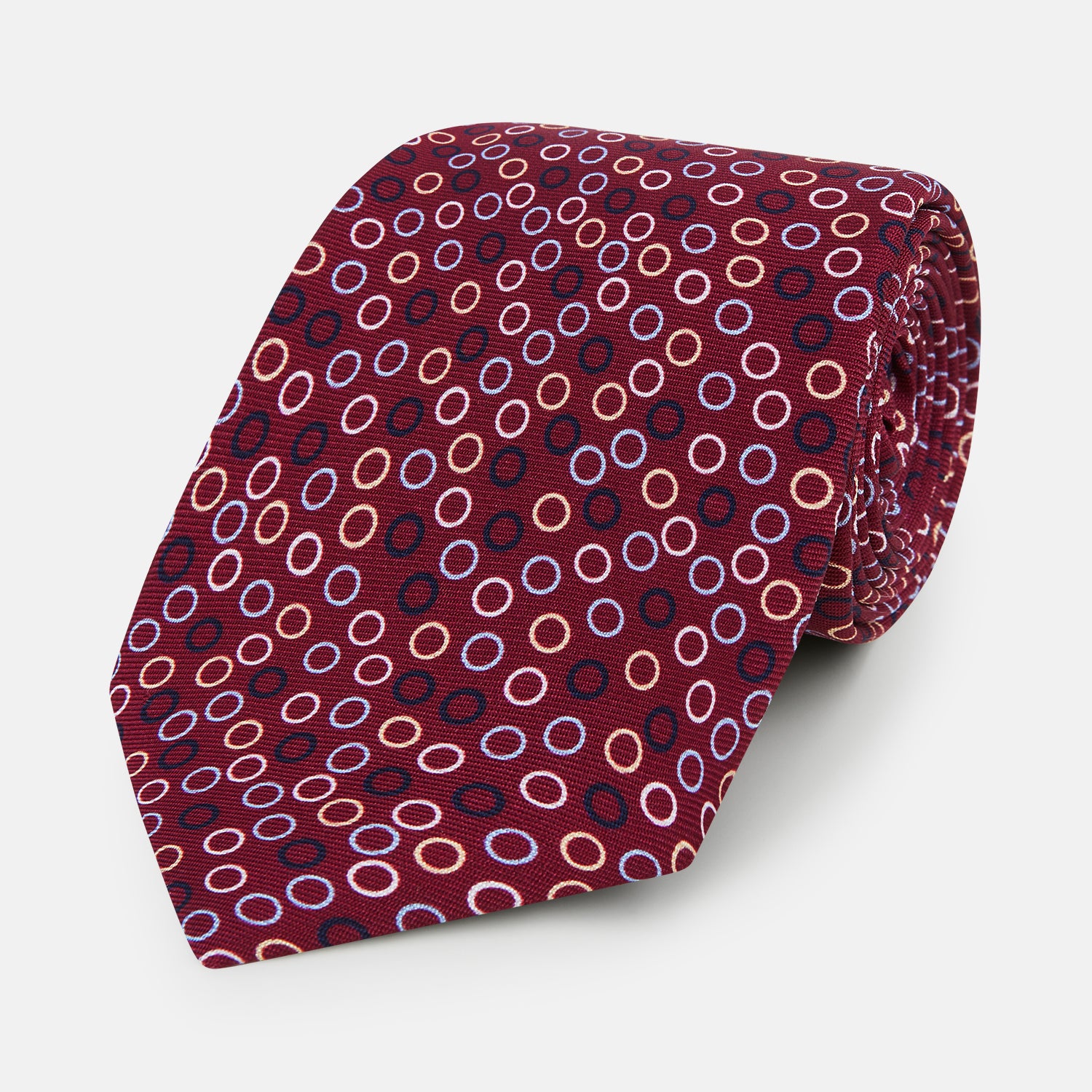 View 1 of product The Turnbull & Asser Pink and Navy Circle Burgundy Printed Silk Tie features overlapping circles in white, light blue, gold, and dark red, presented rolled up on a white background.