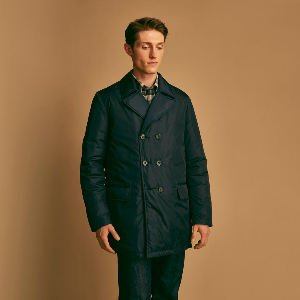 A man stands against a beige background, wearing the Turnbull & Asser Navy Burrows Double Breasted Coat over a plaid shirt and mauve cardigan, paired with dark rolled-up jeans and black shoes, looking directly at the camera with a neutral expression.