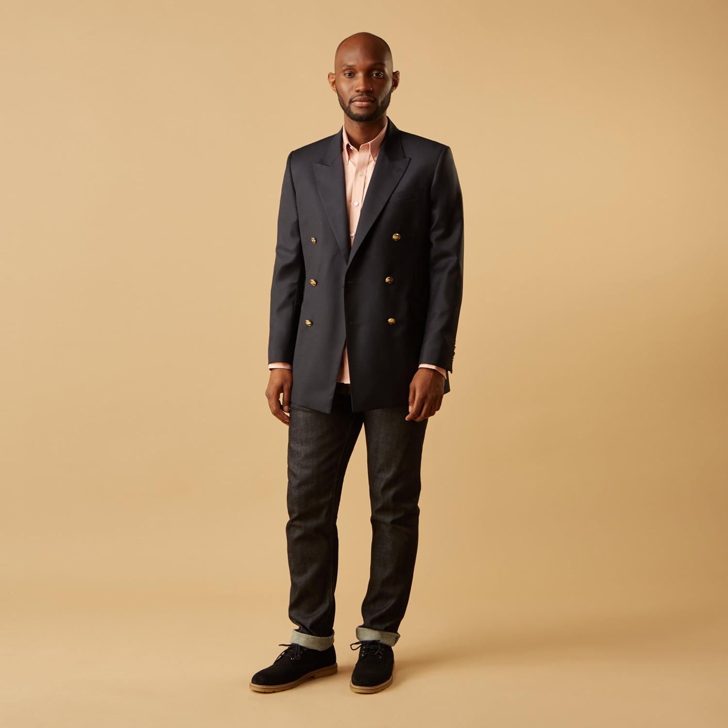 View 7 of product A man faces forward in a relaxed pose against a beige background, wearing the Turnbull & Asser Pink Piece Dyed Weekend Fit Suffolk Shirt with a button-down collar, dark cuffed jeans, and black shoes.