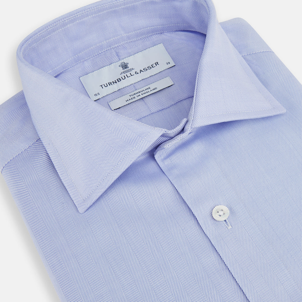 The Turnbull & Asser BLUE HERRINGBONE TAILORED FIT SHIRT WITH KENT COLLAR features long sleeves and buttoned cuffs, offering a timeless formal look in a classic light blue herringbone fabric, laid flat on a white background.