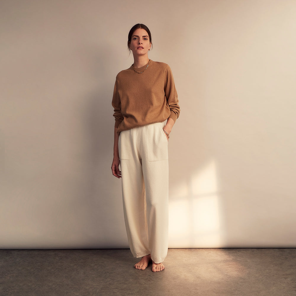 A woman stands barefoot indoors against a neutral wall, wearing Turnbull & Asser Women's Cream Cashmere Knitted Tabitha Bottoms and a light brown sweater. Soft sunlight casts a gentle shadow on the wall beside her.